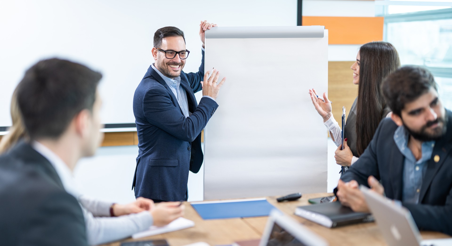 Jitterous businessman stands excitedly next to a clipboard, presenting his ideas to his business colleagues in the meeting room.